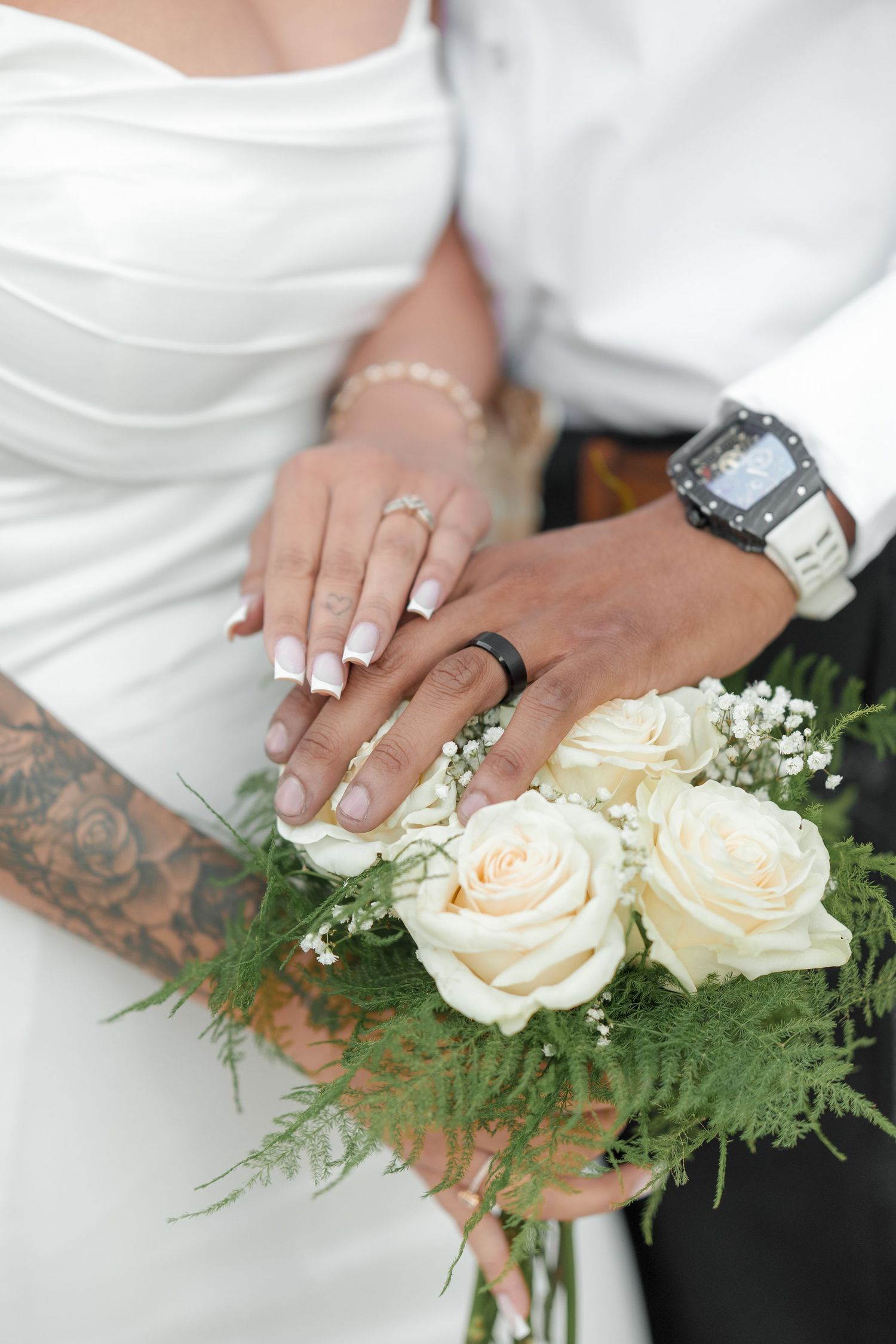 Close-up of tattooed hands holding white roses and baby's breath bouquet with greenery accents.