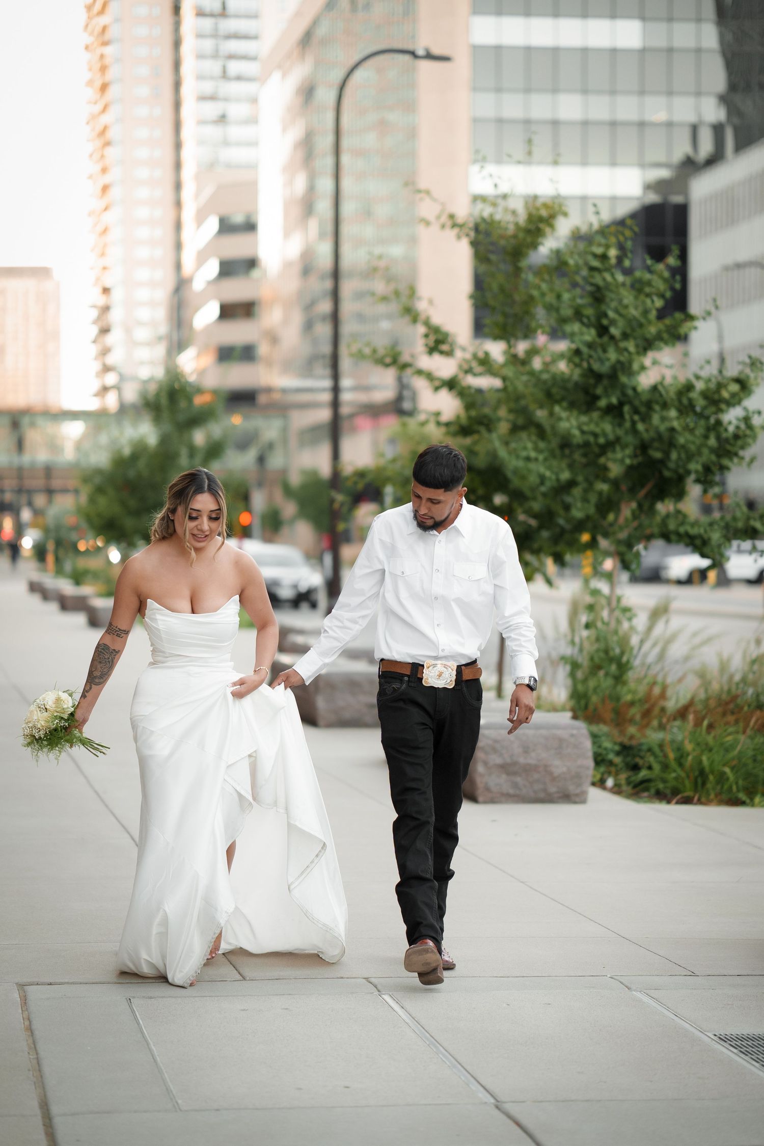 Newlywed couple walking and playing together on an urban sidewalk with city buildings in background.