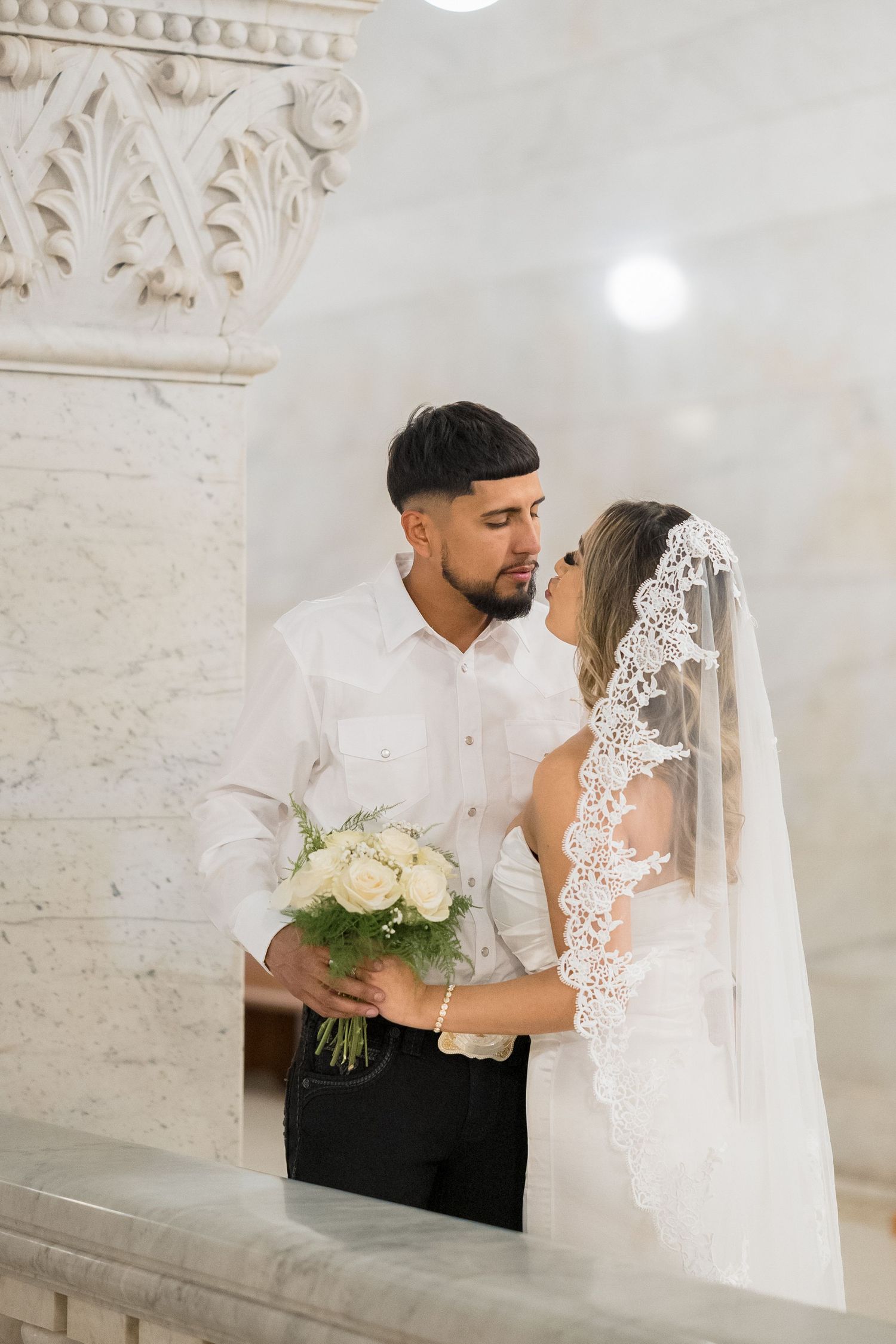 A romantic wedding couple embraces in an elegant white brick venue with ornate architectural details.