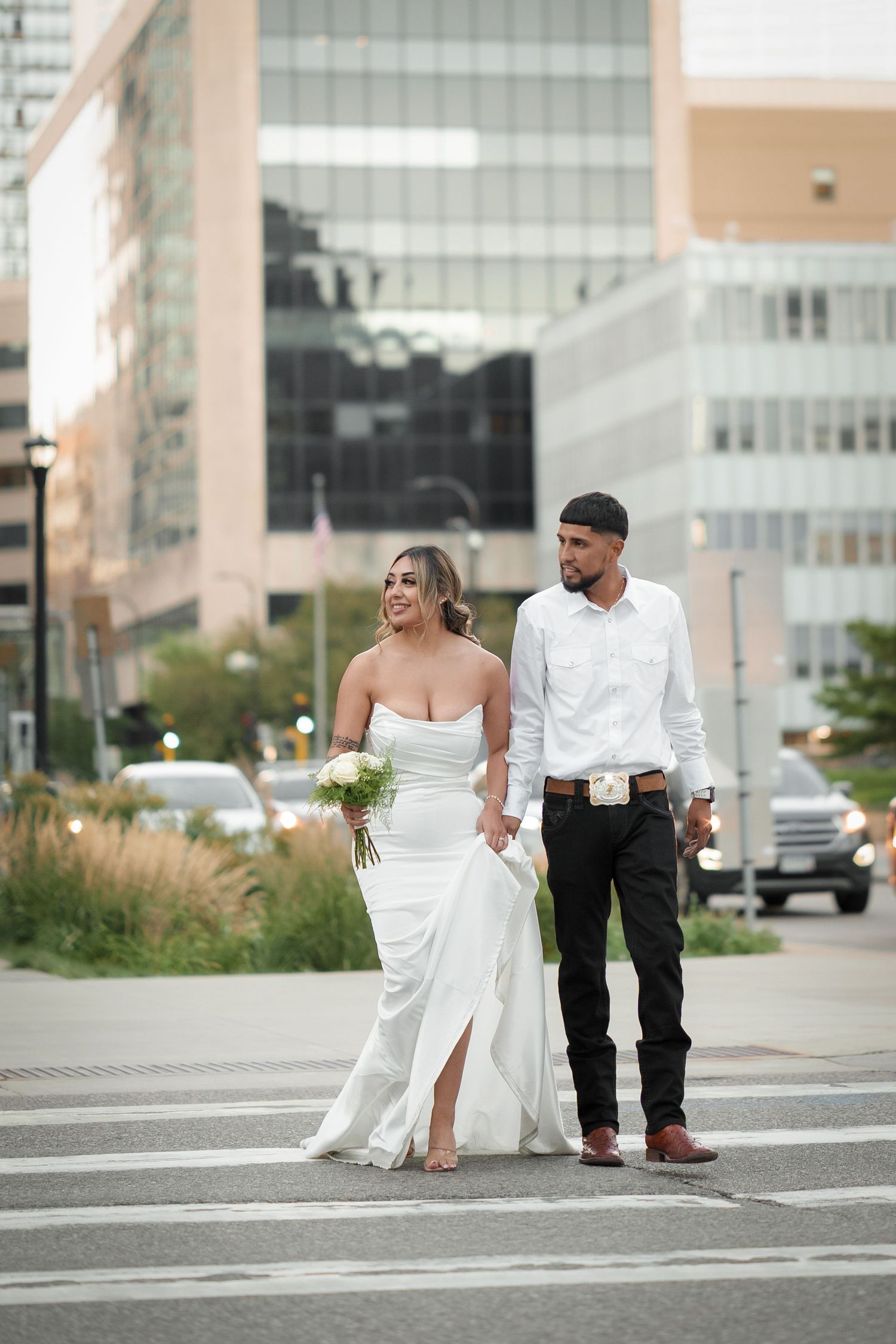 A couple in wedding attire playfully walks across a crosswalk in an urban setting.