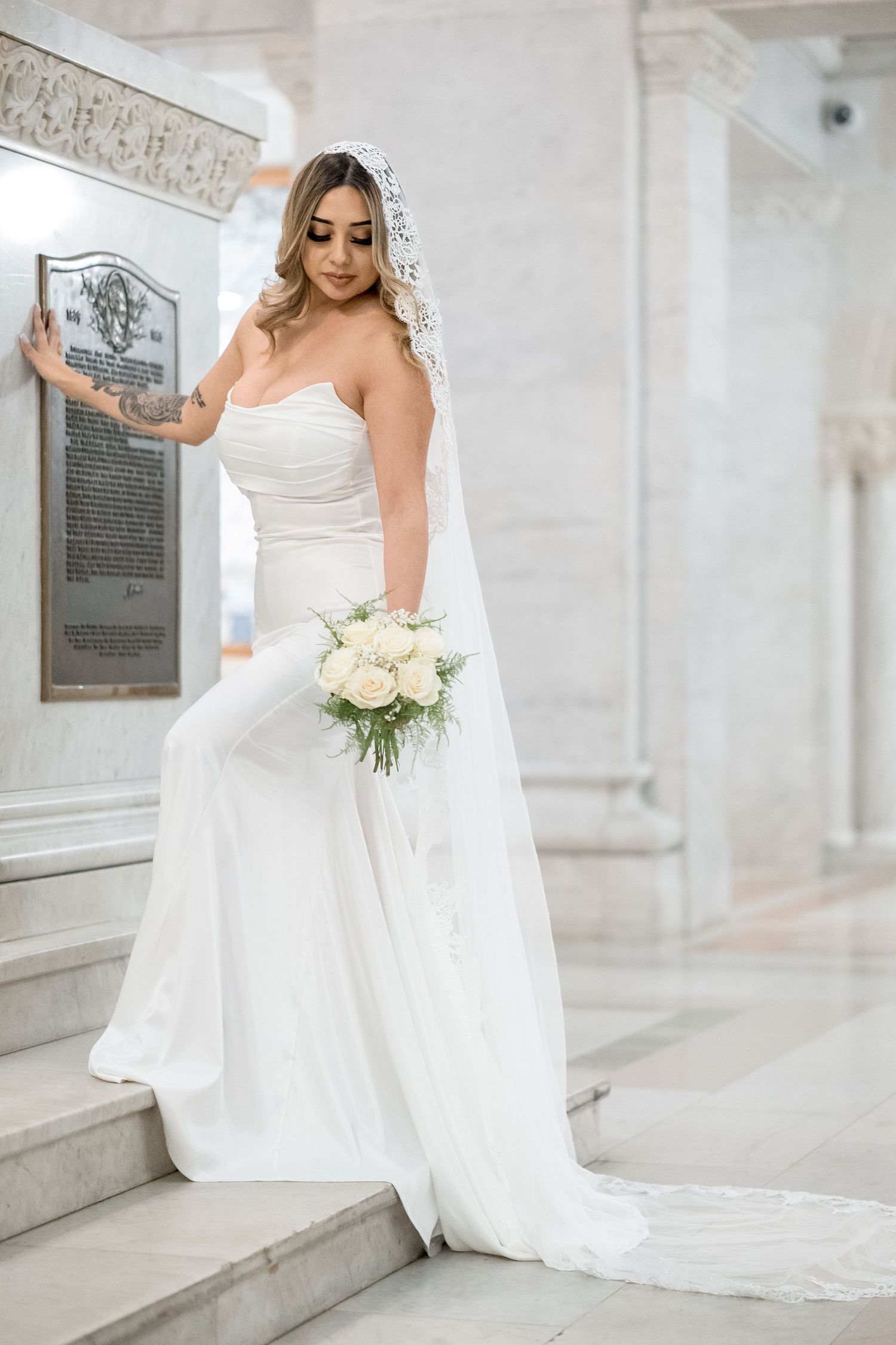 A bride in a white wedding gown and veil poses elegantly in a light-filled marble hallway.