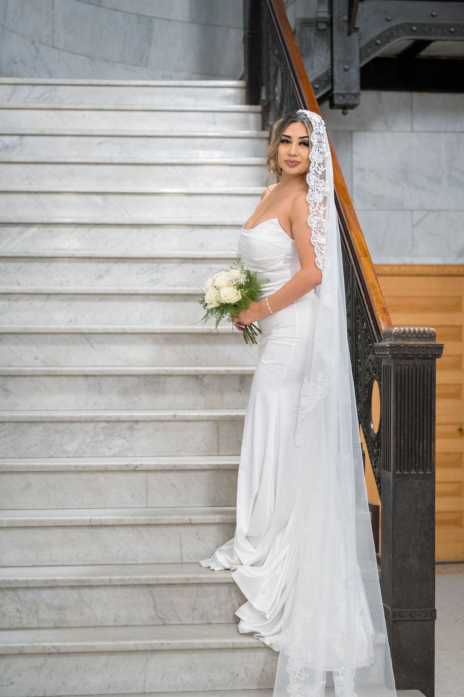 A bride in a white wedding dress poses gracefully on marble stairs with a wooden banister and holds a white bouquet.