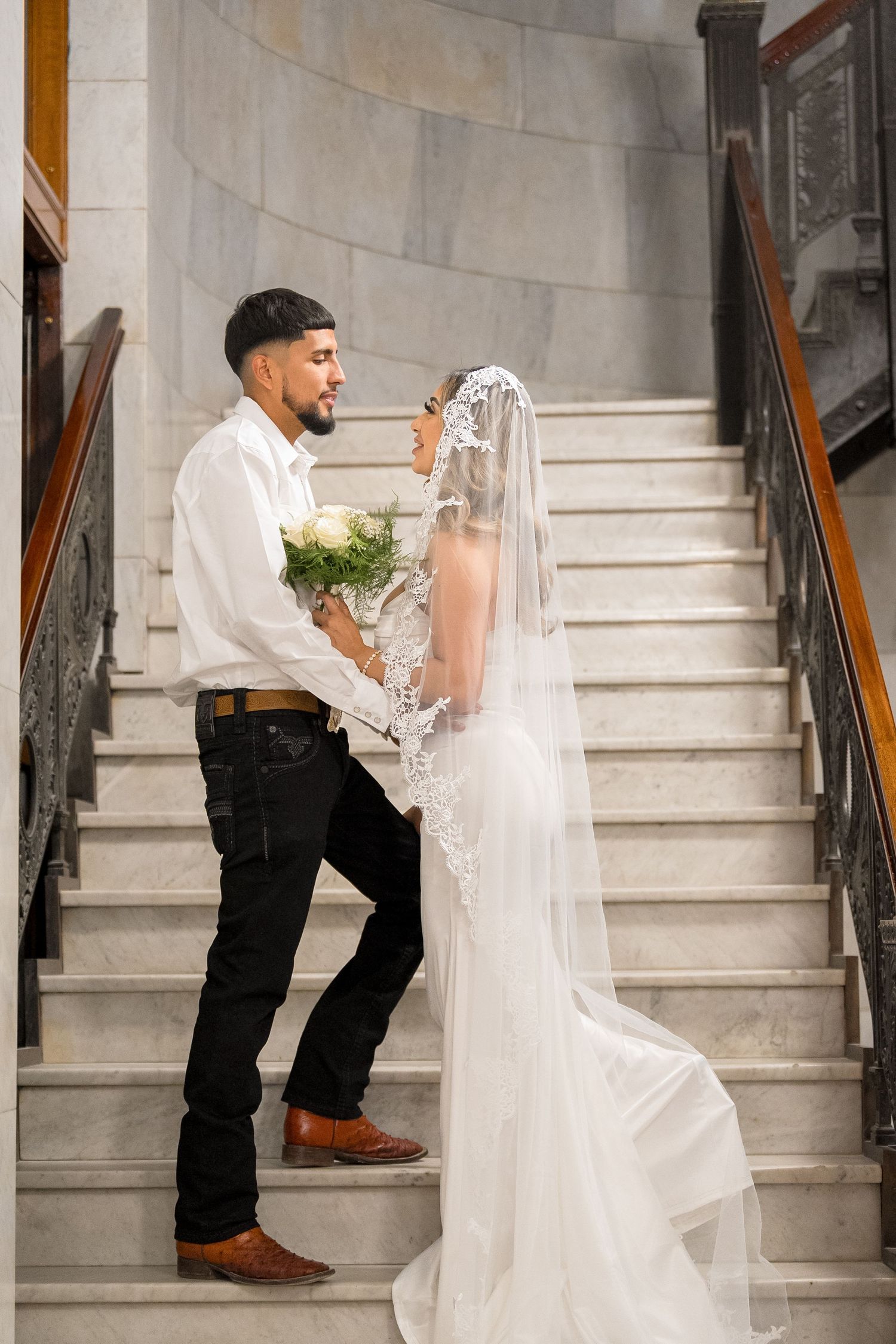 A bride in a flowing white dress and veil descends elegant marble stairs while holding a bouquet.