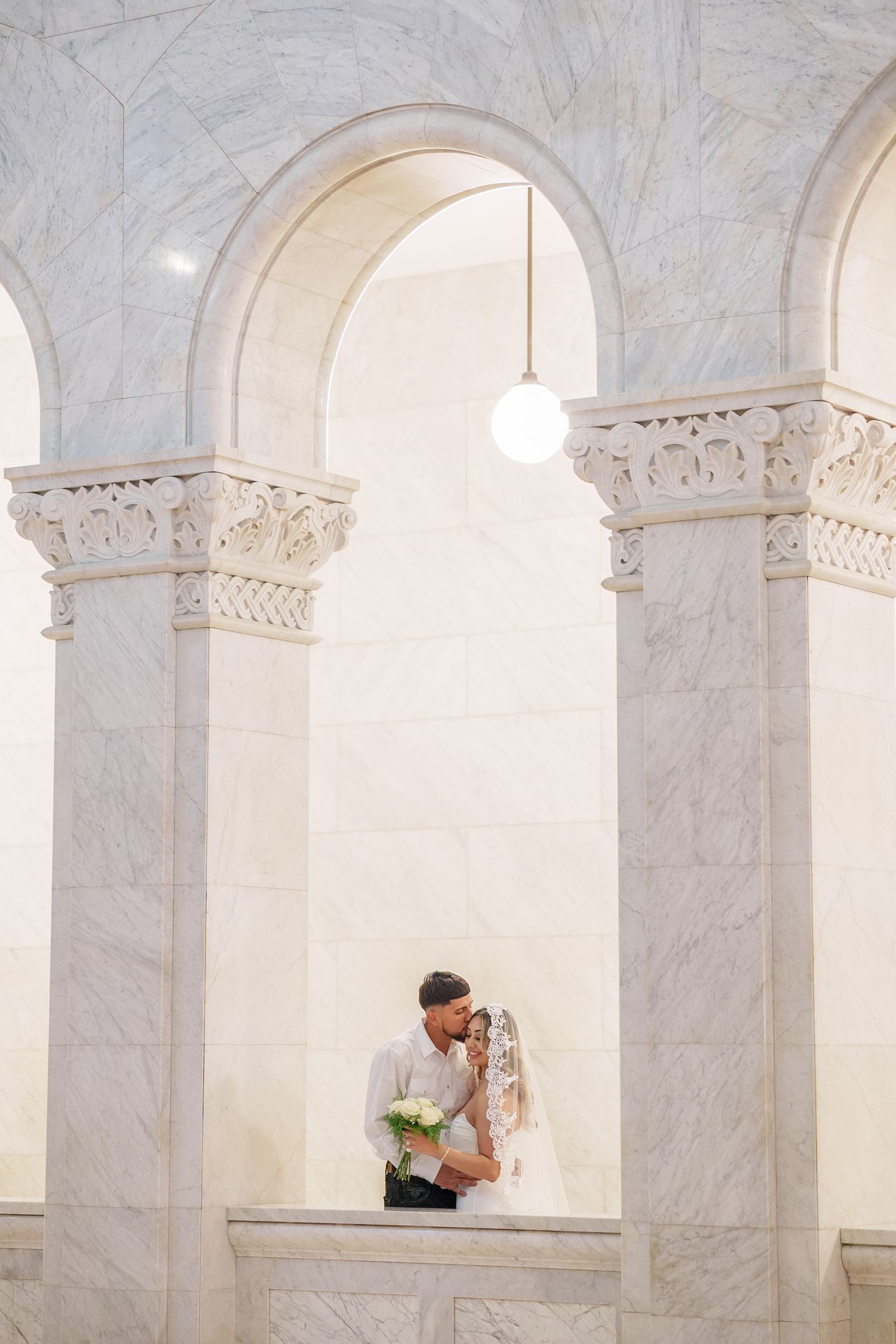 A couple shares a romantic moment under elegant white marble arches in a classic architectural setting.