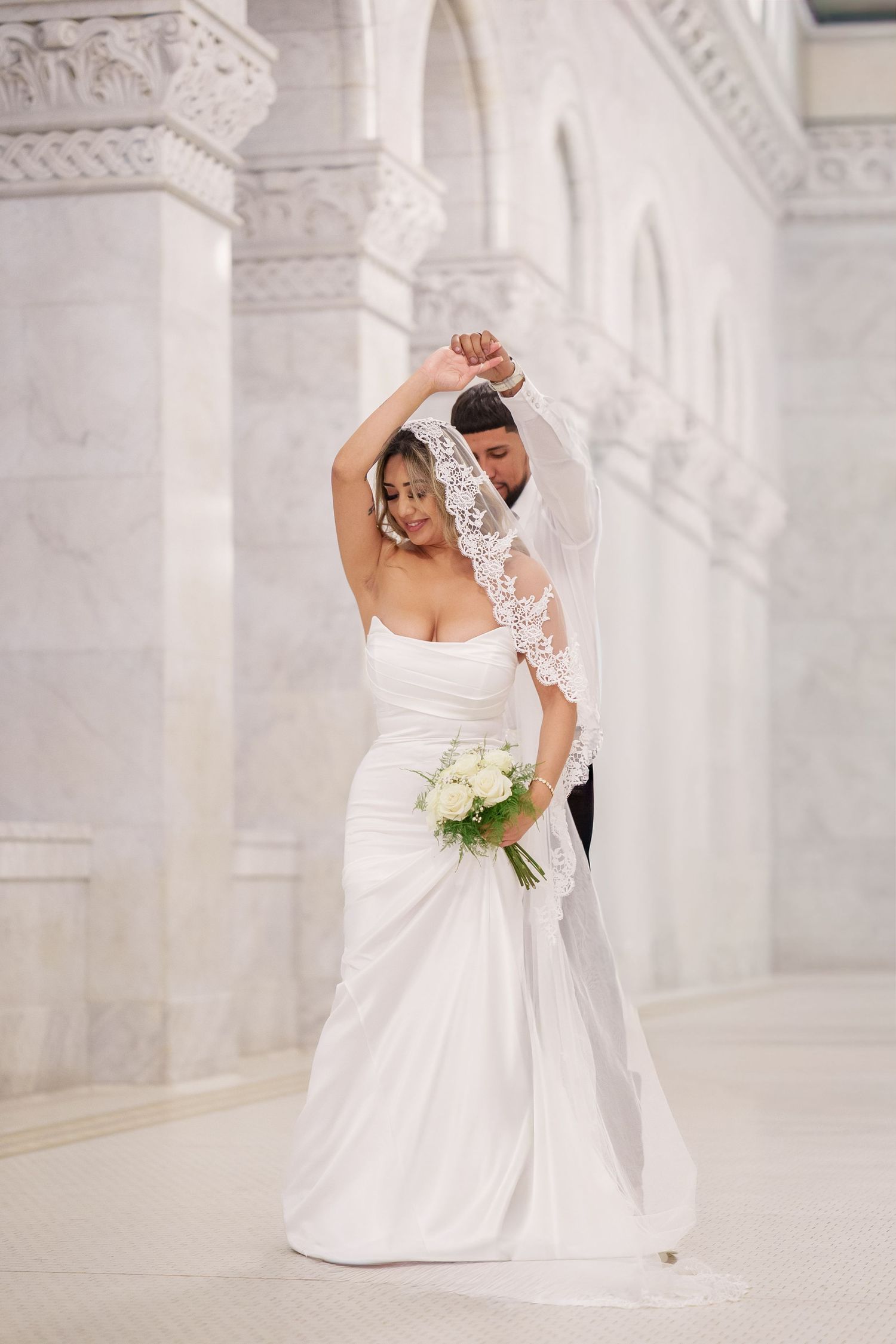 Elegant bride in white strapless gown with lace veil poses gracefully against ornate white stone archways.