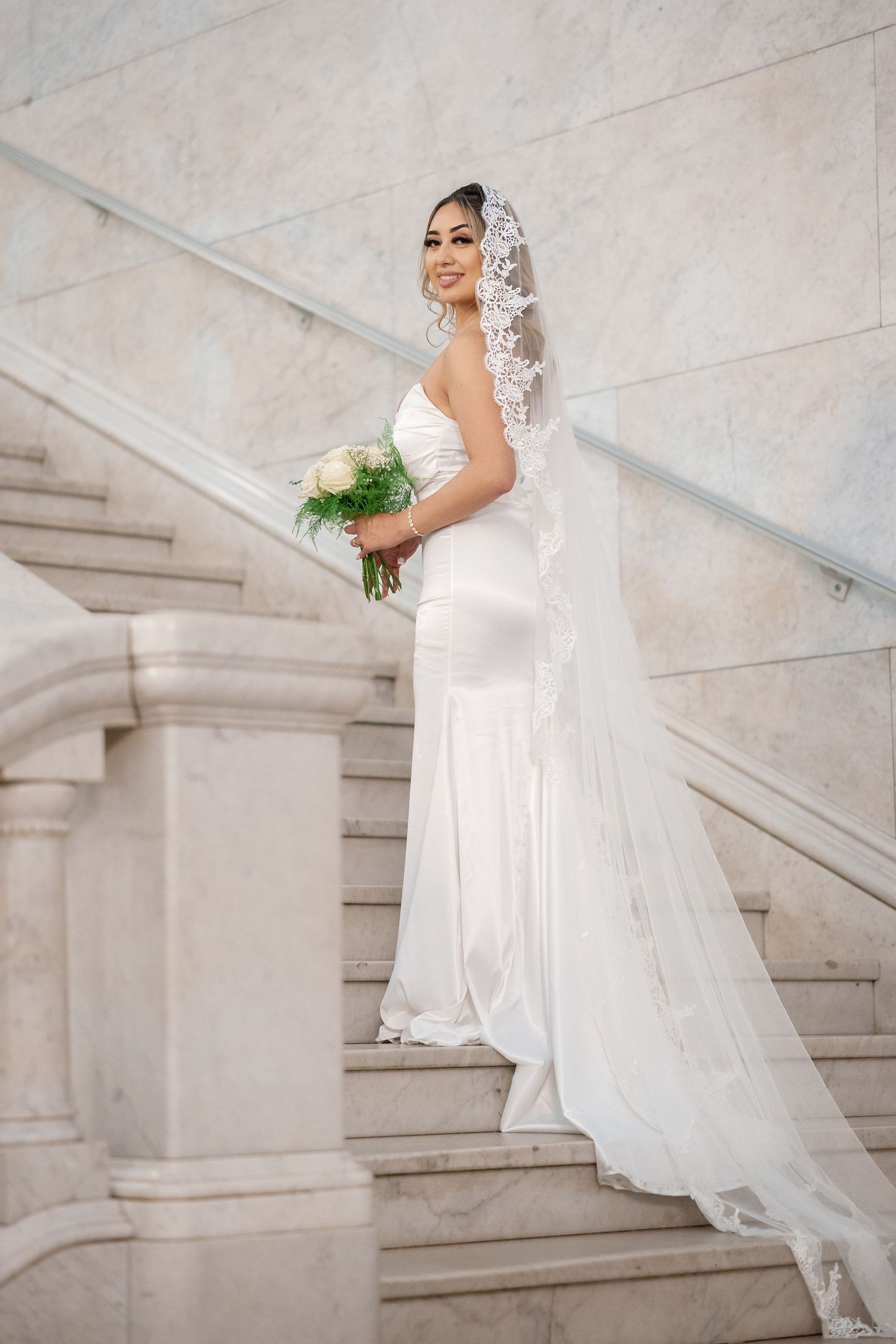 A bride in an elegant white wedding dress and lace veil poses on marble stairs while holding a bouquet.