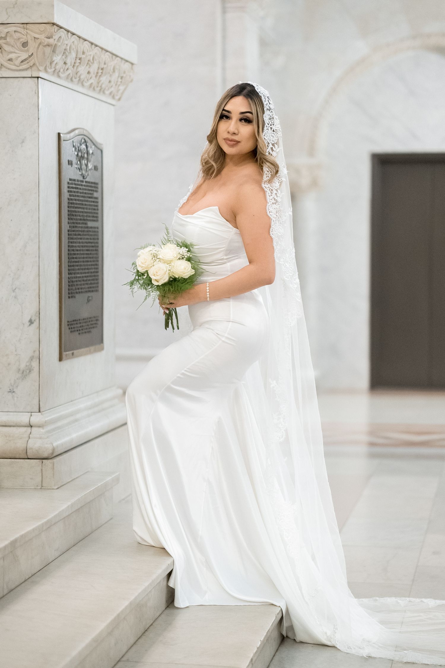 A bride in a strapless white wedding gown and veil holds a bouquet of white roses while standing on marble steps.