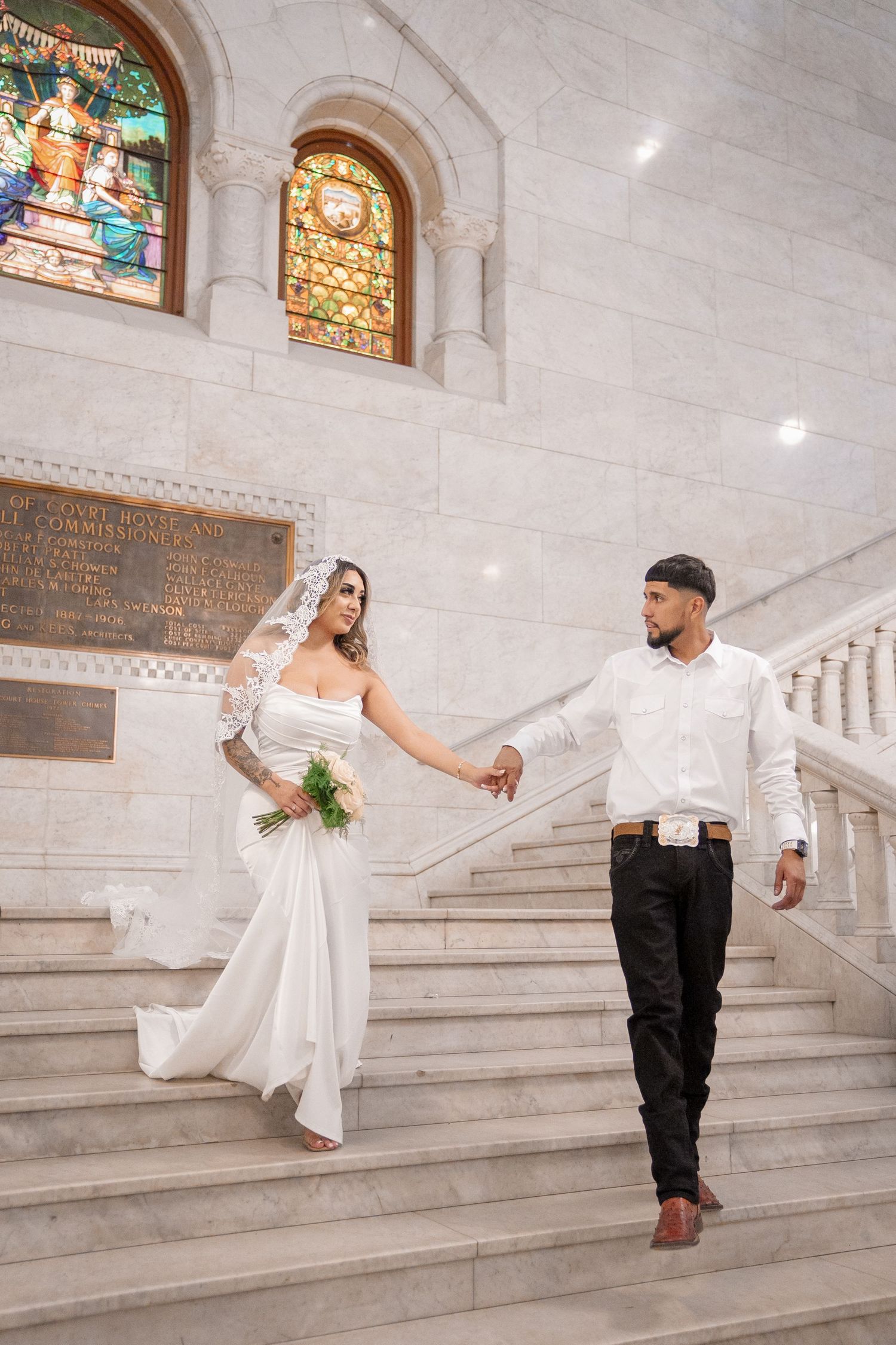 A couple poses playfully on elegant marble stairs with stained glass windows in the background.