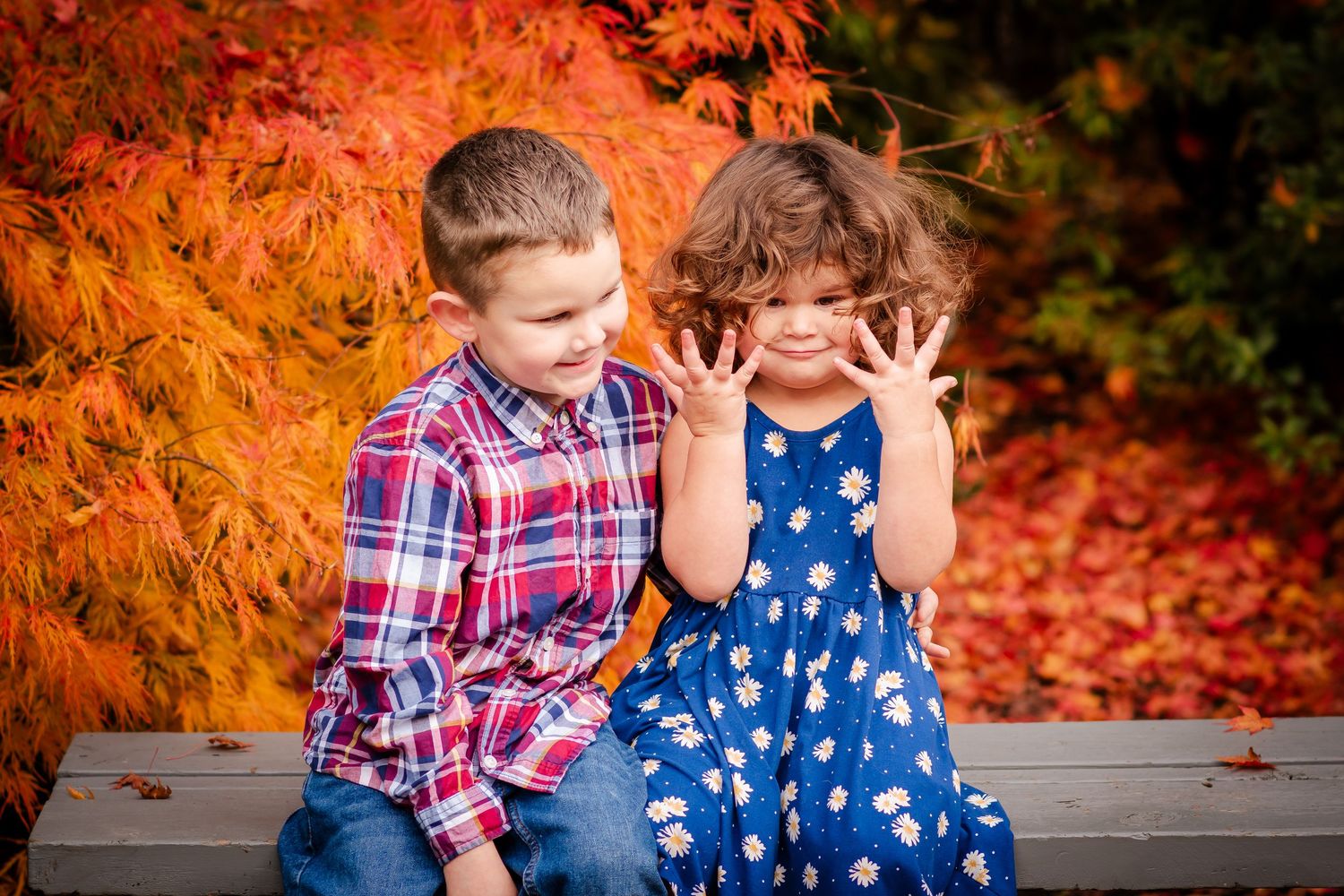Two children sit together on a bench against vibrant orange autumn foliage in the background.
