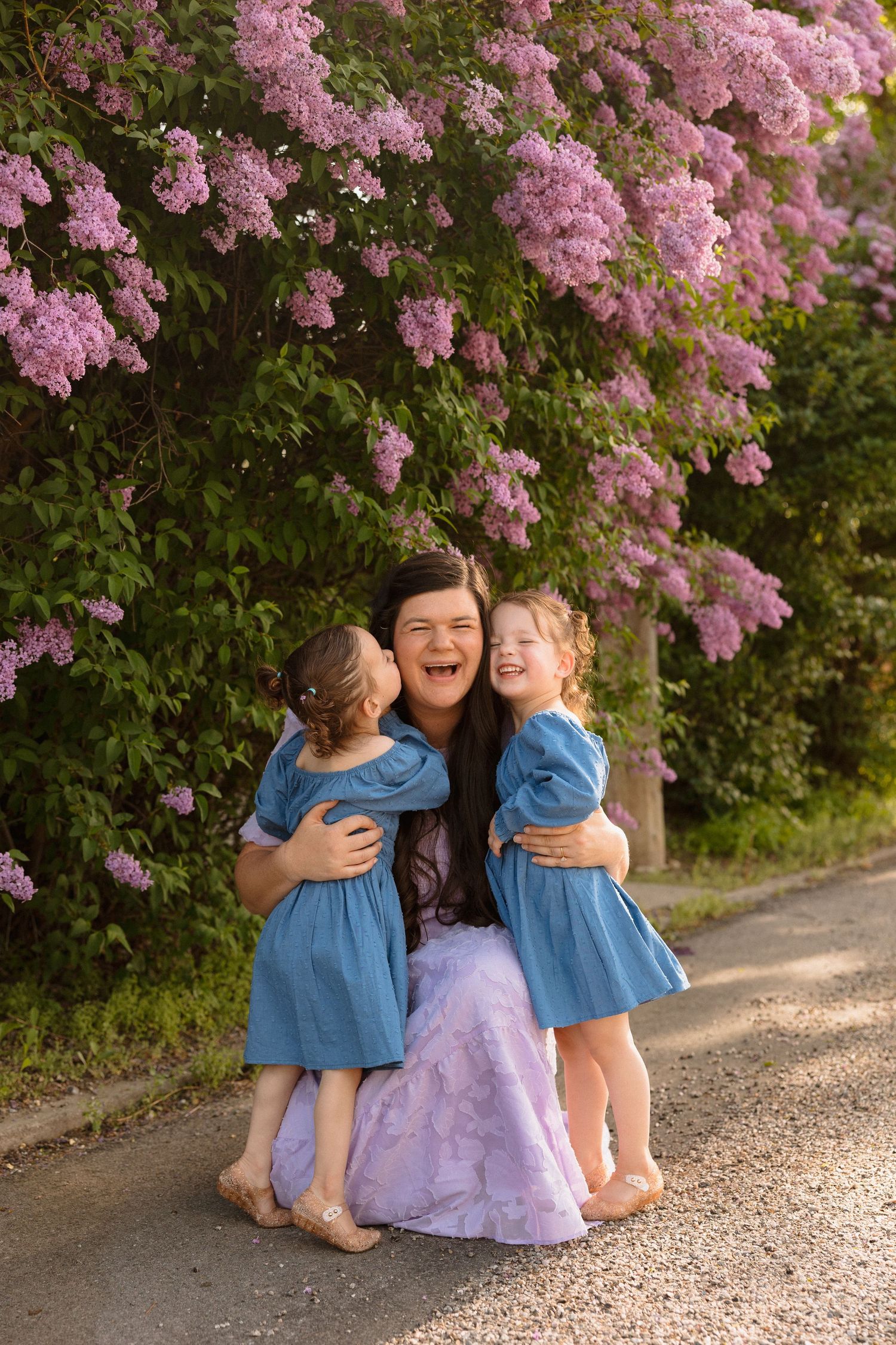 A joyful family embrace under blooming pink lilac bushes on a gravel path.