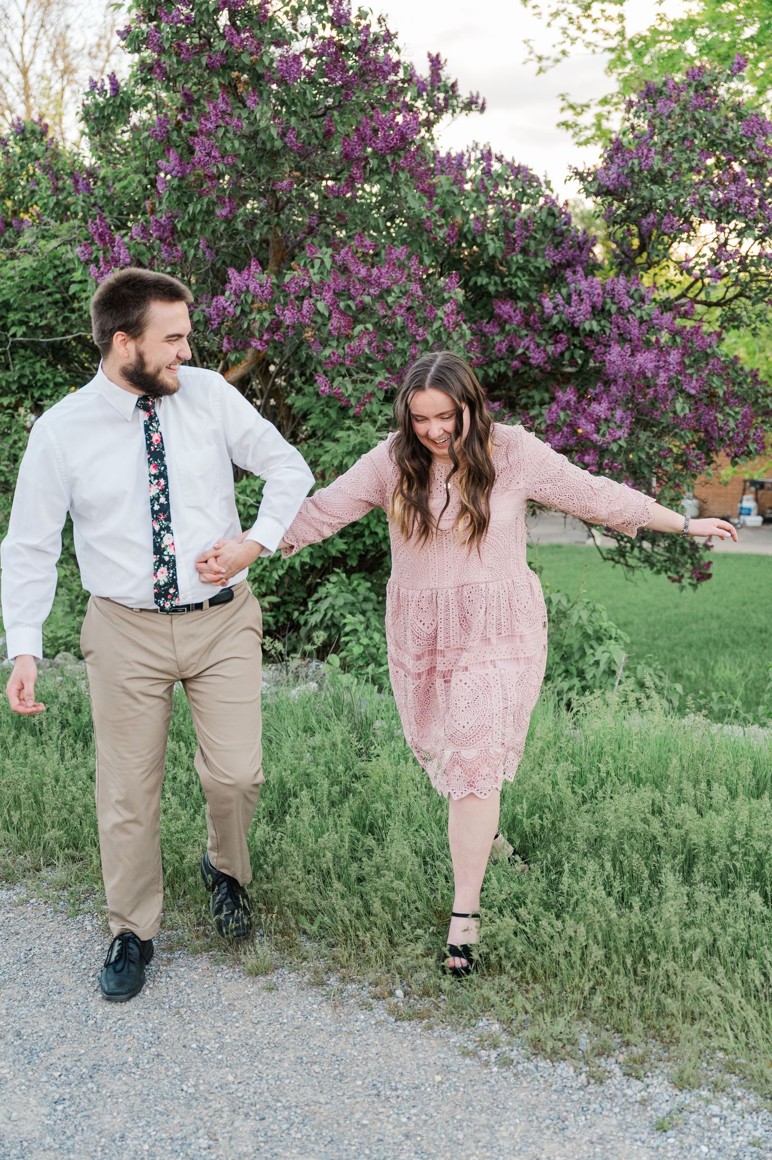 Couple holding hands and laughing together on a path with blooming lilac bushes in the background.