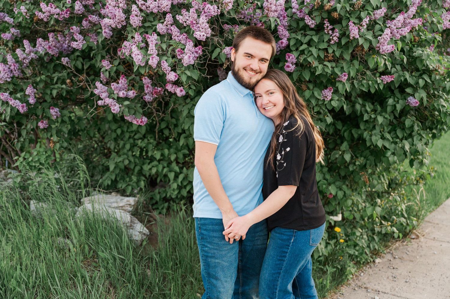 Couple embracing in front of purple flowering lilac bush.