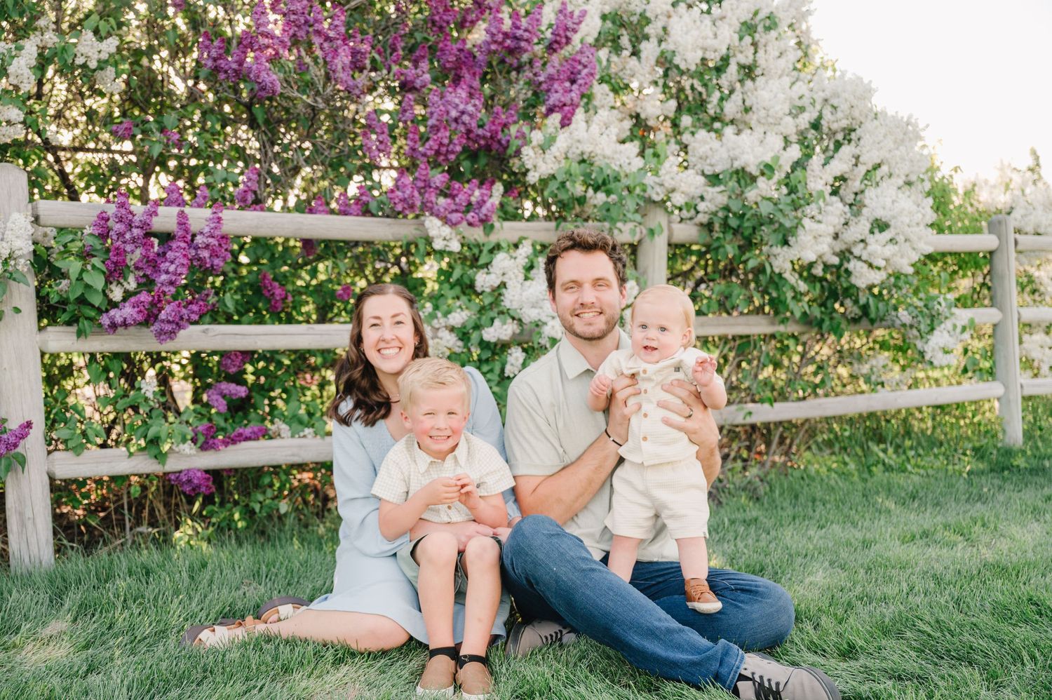 A family sits on a wooden bench outdoors with blooming purple bougainvillea flowers and green foliage in the background.