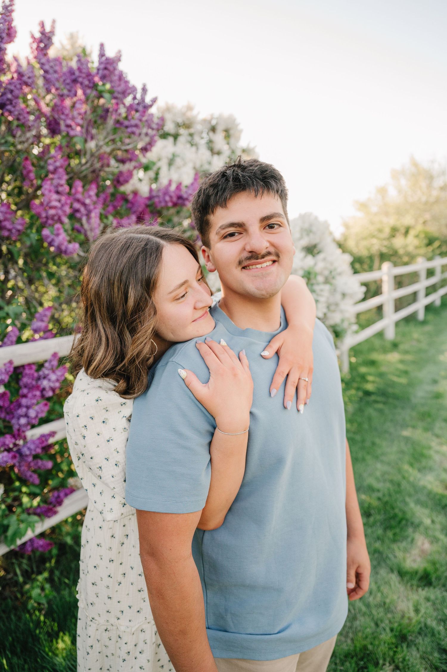 A couple embrace by blooming purple flowers and a white fence in an outdoor garden setting.