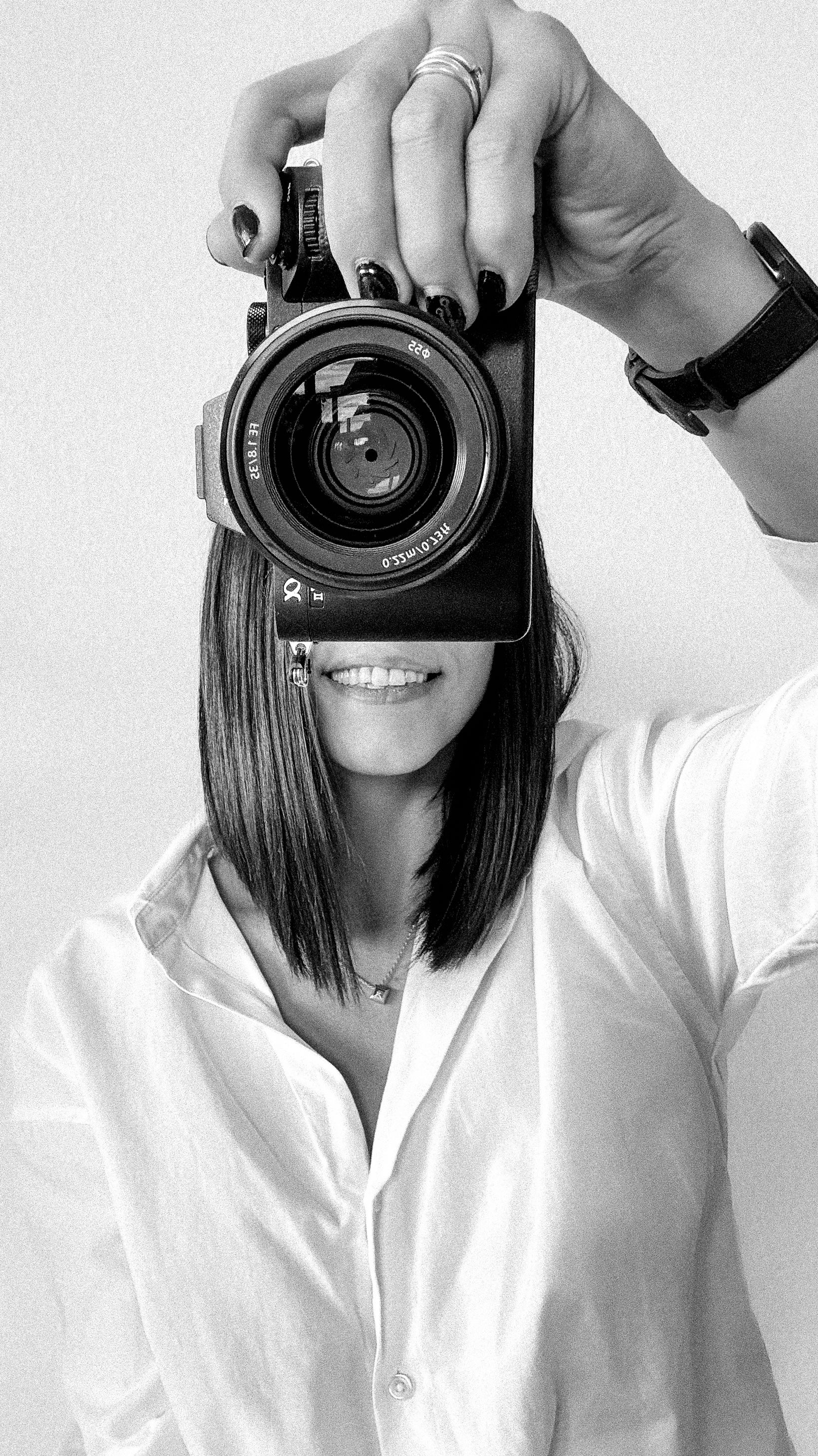 A stylish photographer takes a self-portrait with a professional DSLR camera in black and white.