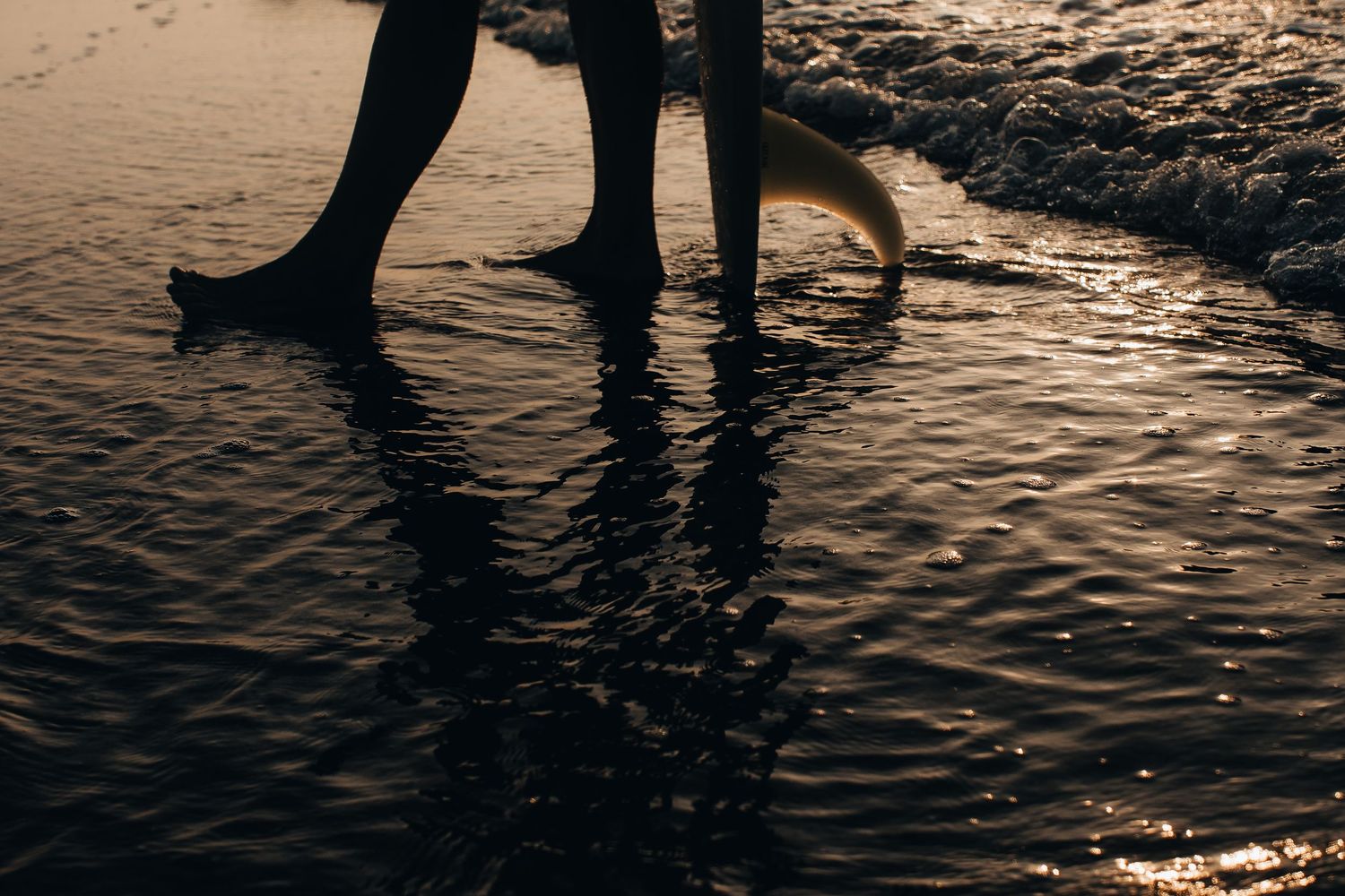 Silhouetted feet walking in shallow water at sunset creating reflections on wet sand.