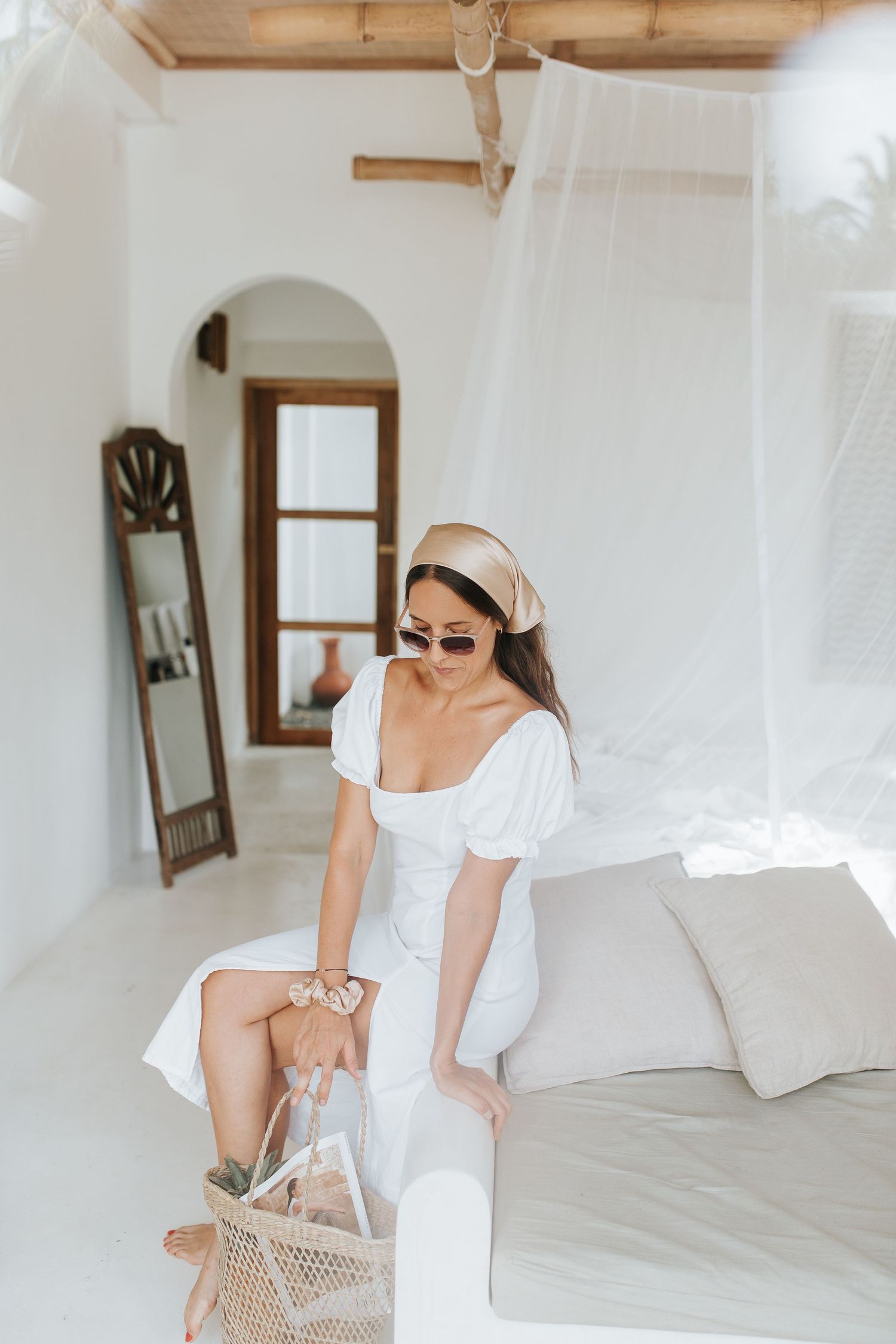 Woman in white dress arranging linens and pillows in a bright, airy bedroom.