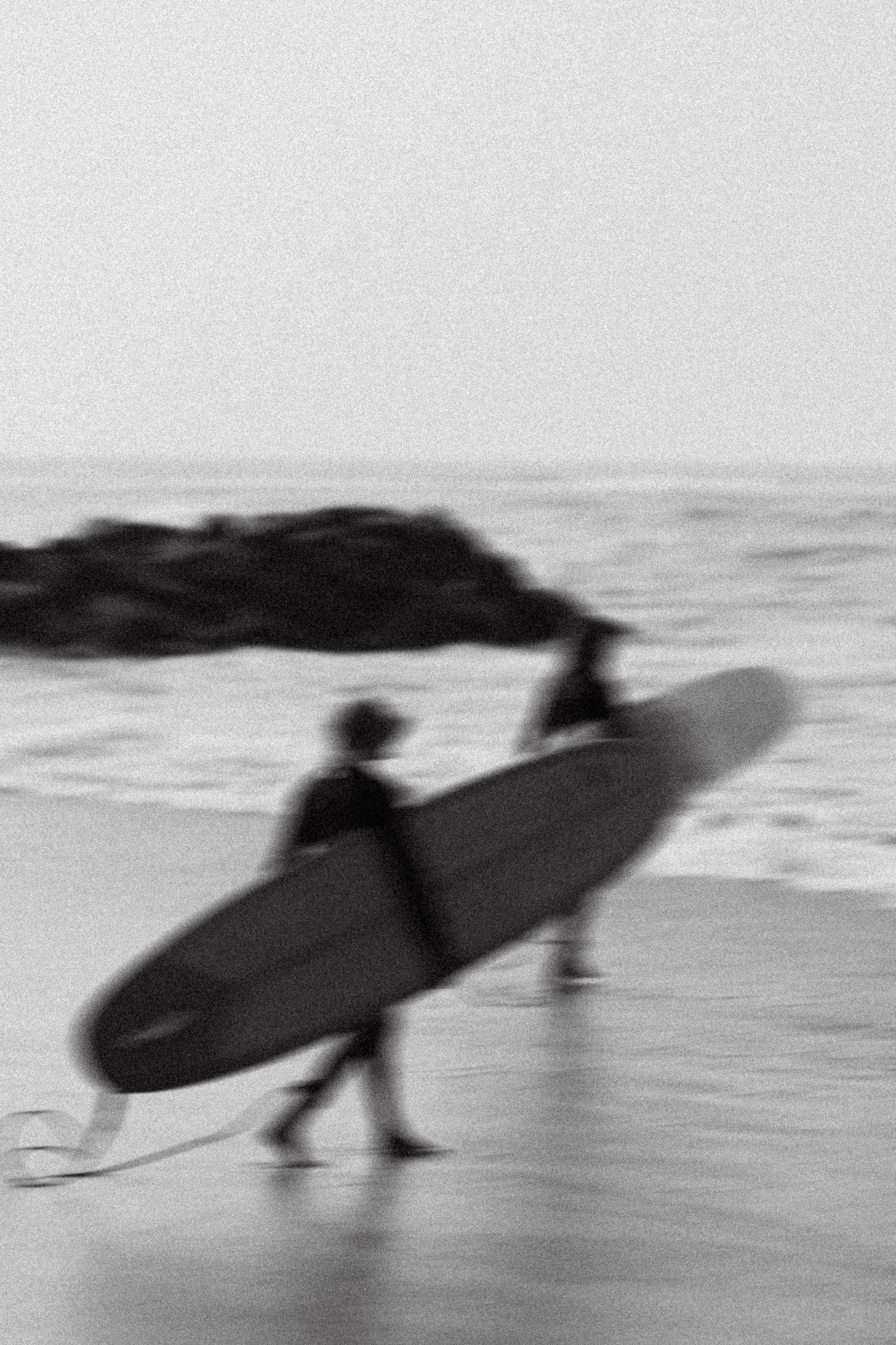 A blurred black and white image of surfers carrying boards on a beach.