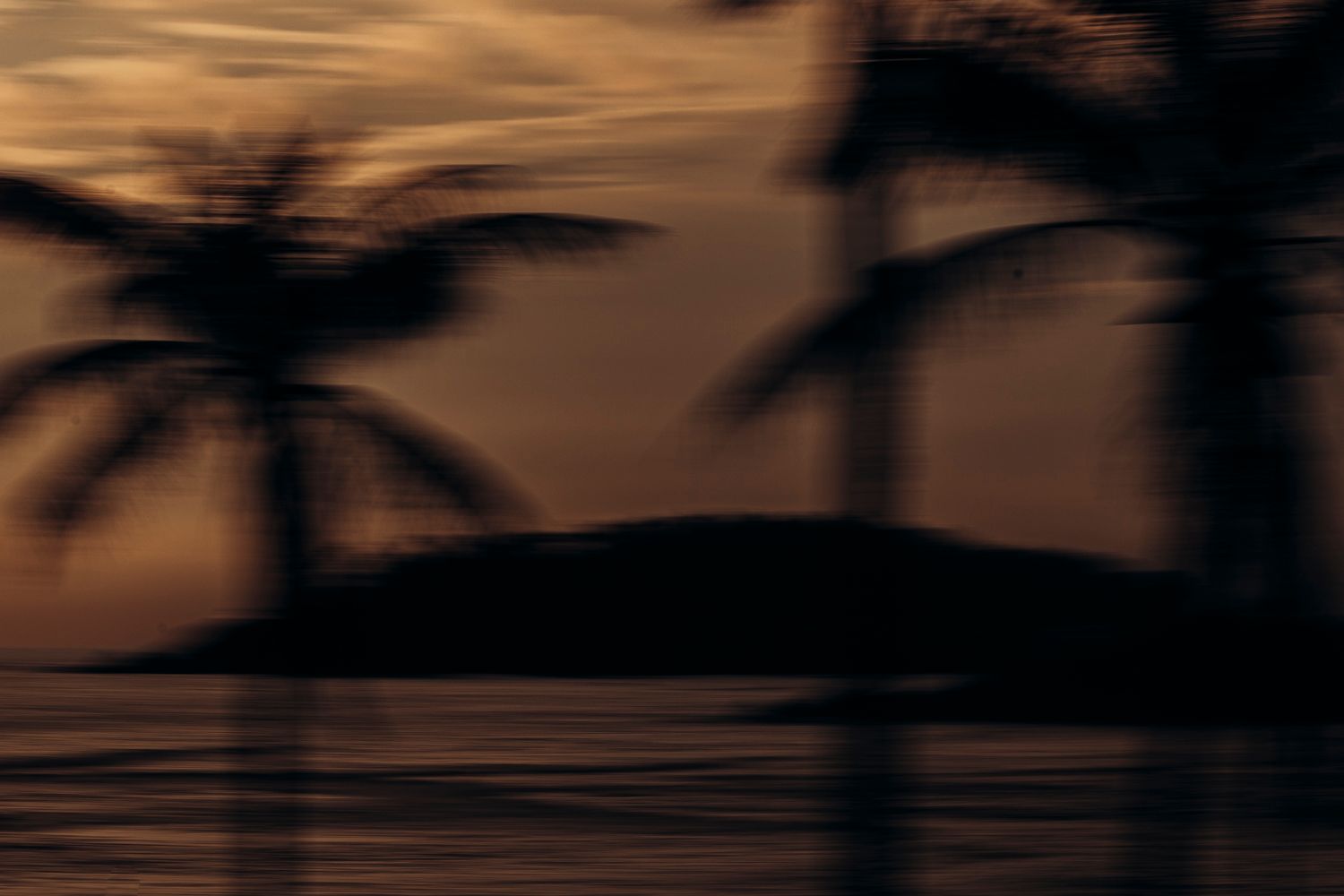 Silhouetted palm trees against a warm orange sunset sky reflecting over calm tropical waters near an island.