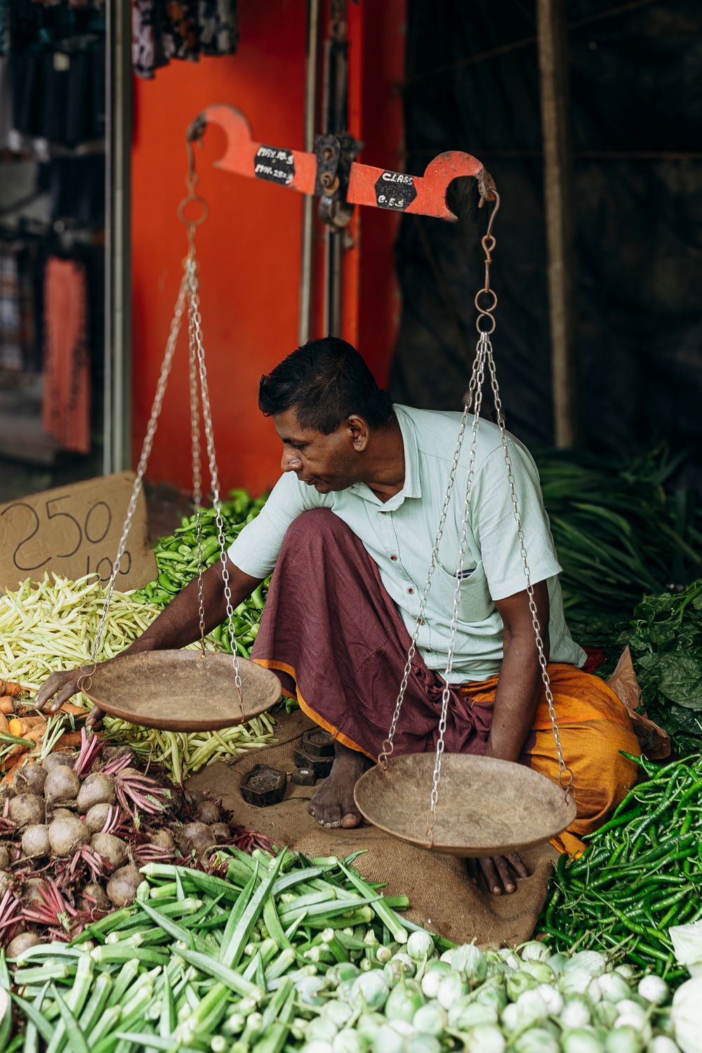 A man sits cross-legged at an outdoor vegetable market stall, using traditional balance scales to weigh fresh produce.