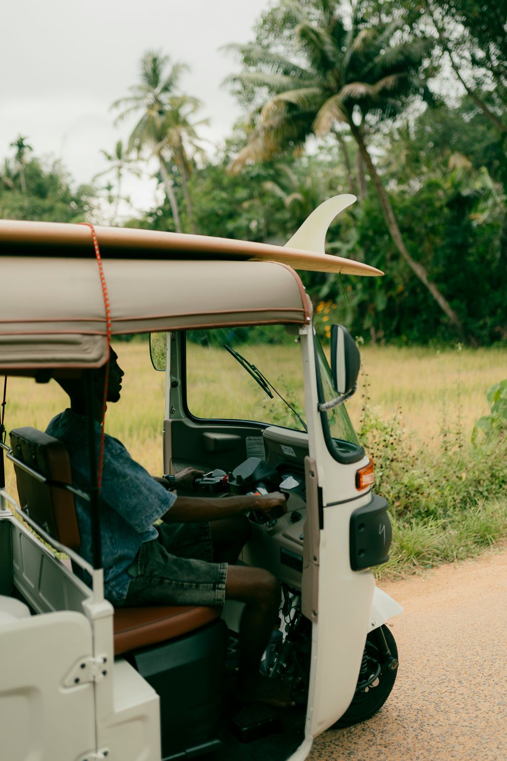 Person driving a tuk-tuk with a surfboard on top through a tropical road surrounded by palm trees.
