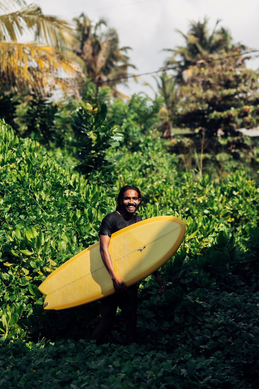 Smiling surfer holding a yellow surfboard among lush tropical greenery and palm trees.