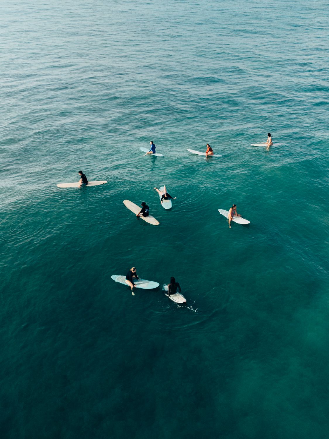 Aerial view of surfers with white surfboards floating on calm turquoise ocean water.