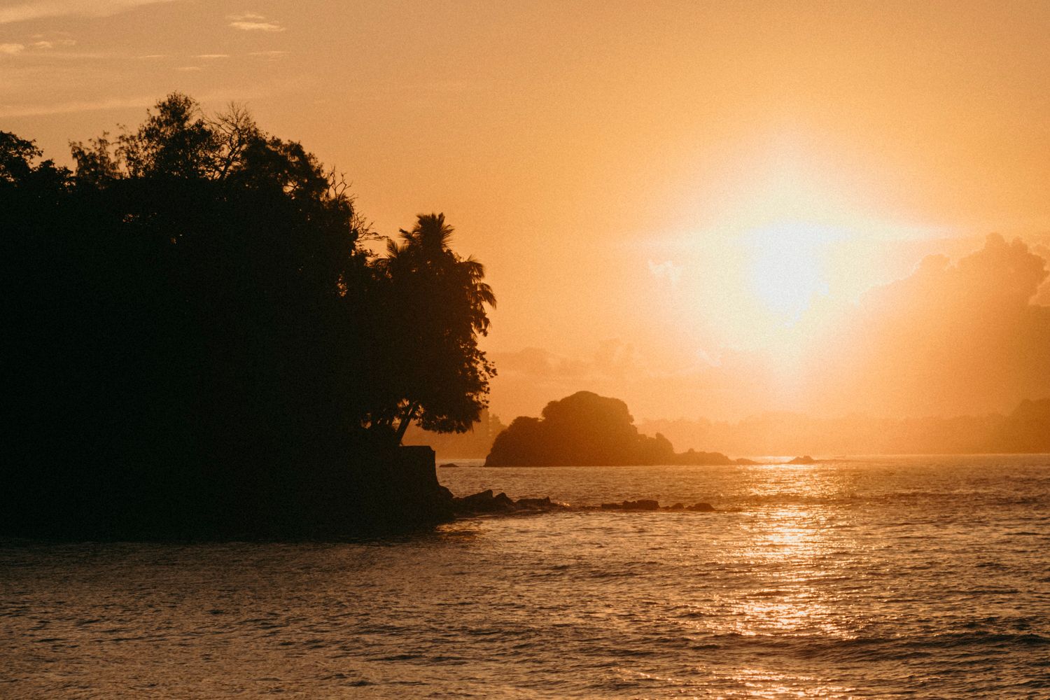 Golden sunset over tropical island silhouette with glowing orange sky reflecting on calm ocean waters.