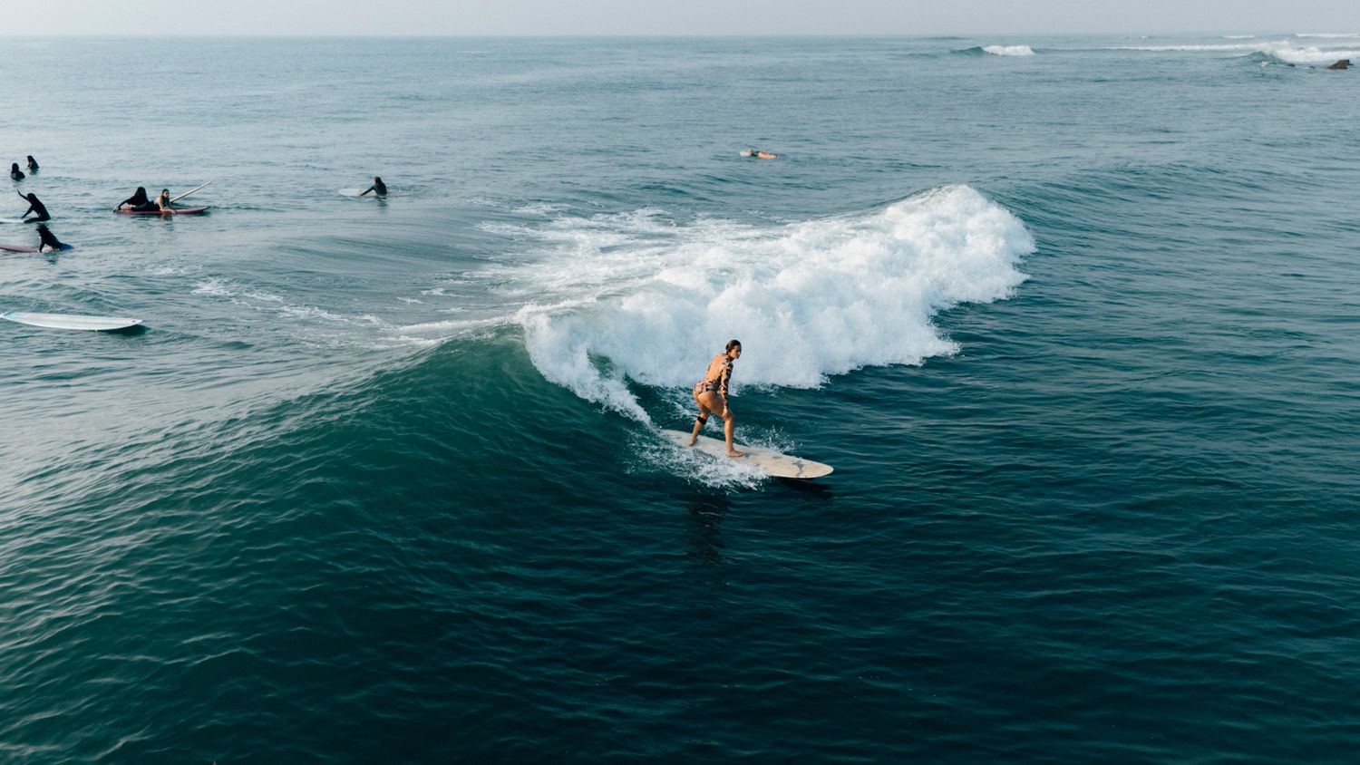 Aerial view of a surfer riding a wave on a white surfboard in the ocean with other surfers in the background.
