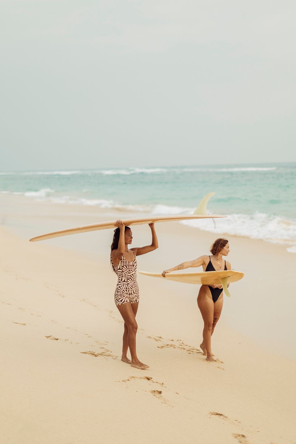 Two women in swimsuits carry yellow surfboards along a sandy beach with turquoise ocean waves in the background.