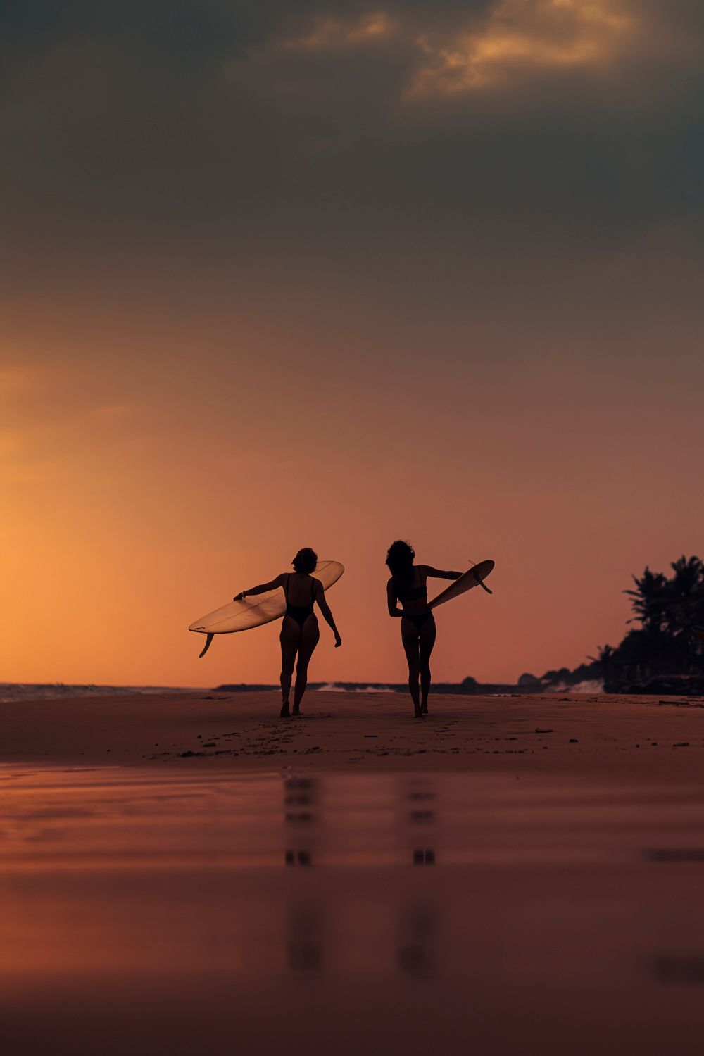 Two surfer silhouettes carry boards on a glowing orange sunset beach, reflecting on wet sand.