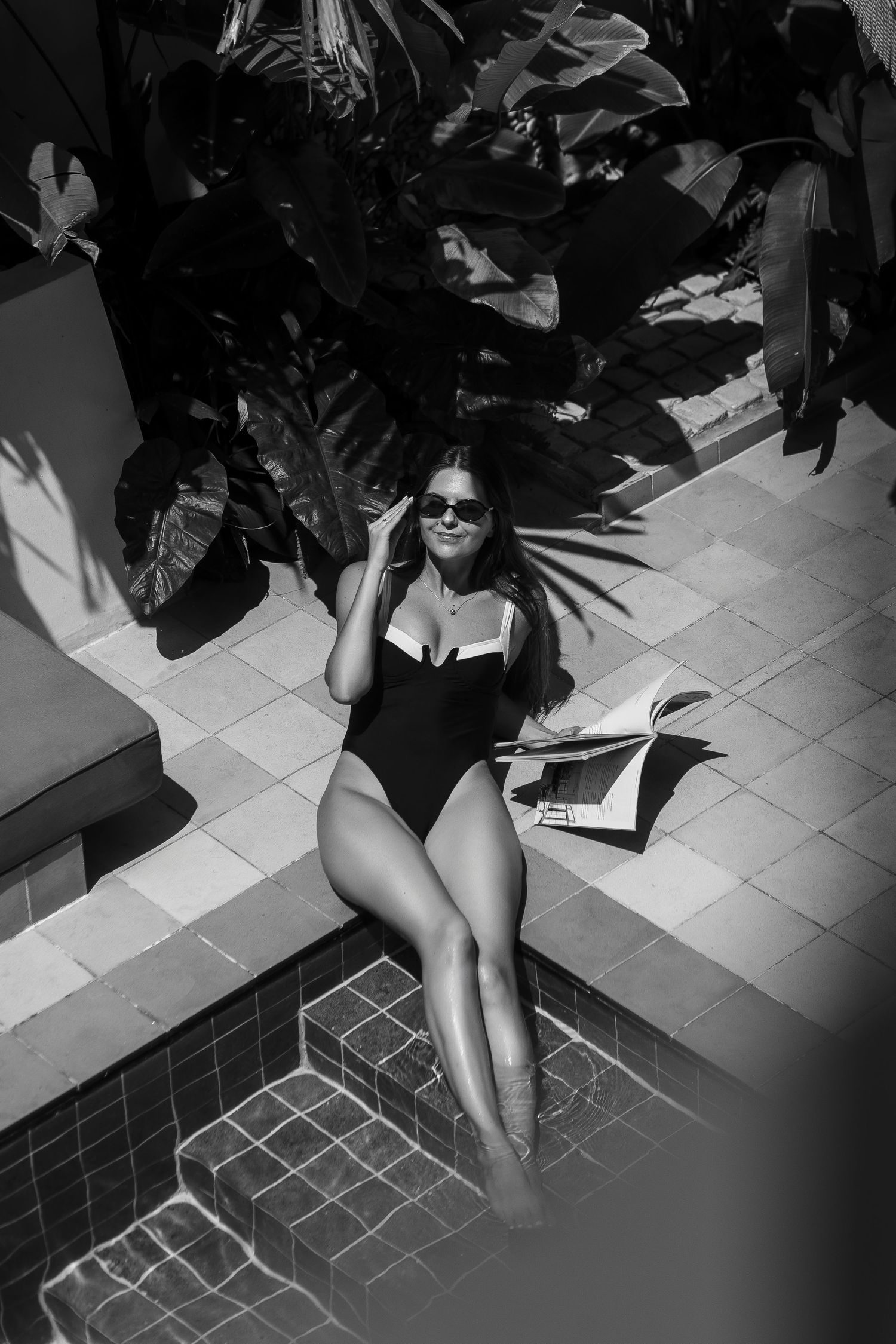 A black and white image of someone lounging by tropical plants on tiled flooring.
