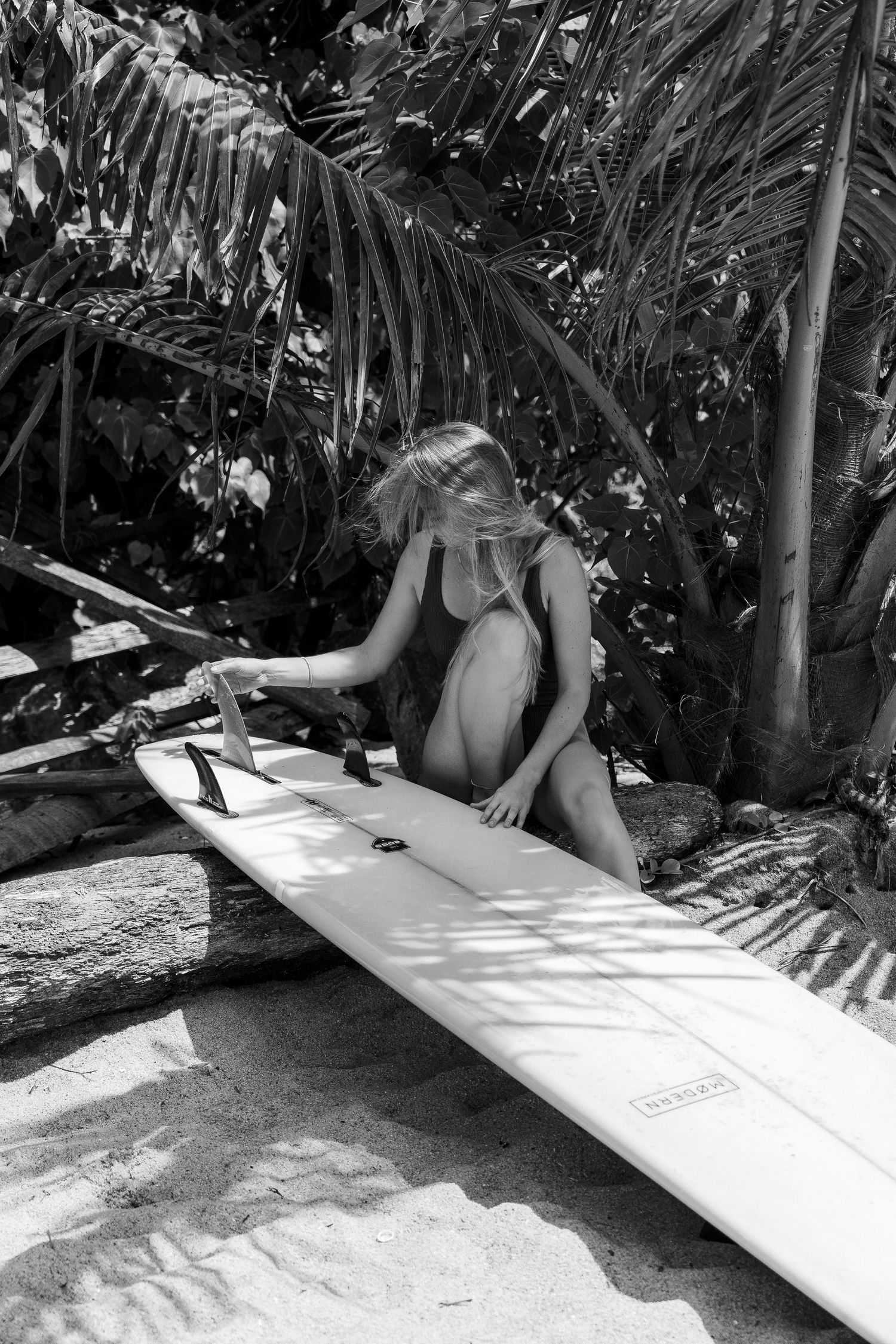 Surfer waxing and preparing a longboard surfboard in a tropical garden setting.