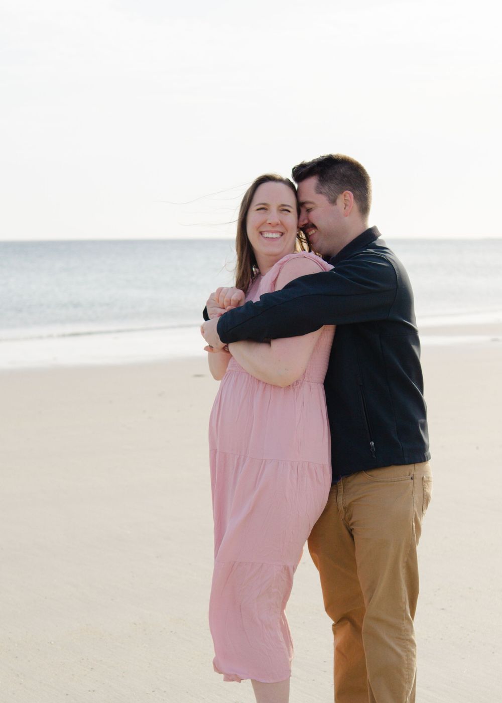 Couple embracing and laughing together on a sunny Cape Cod beach in casual outfit.