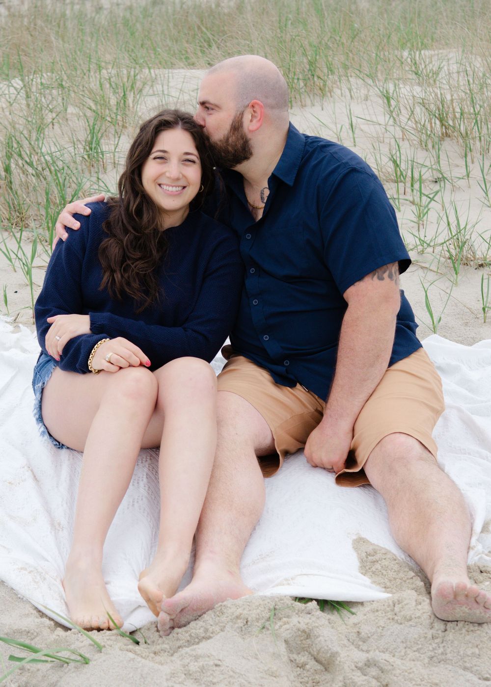 Light-filled beach session, pair sitting among tall grass, enjoying quiet connection.