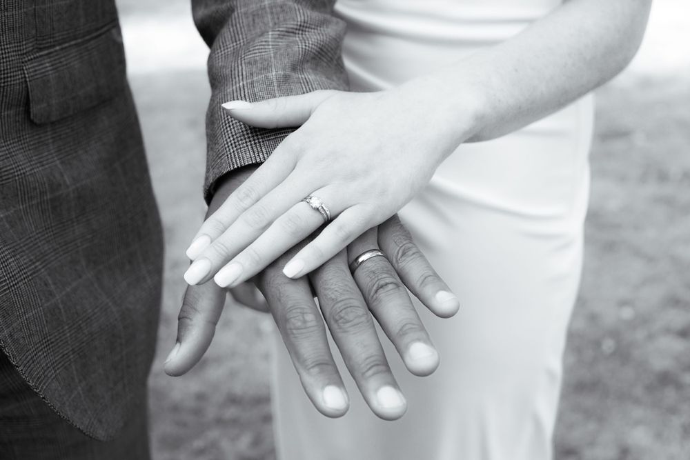 Detail shot, newlyweds’ hands with wedding bands, elegant clothing in background.