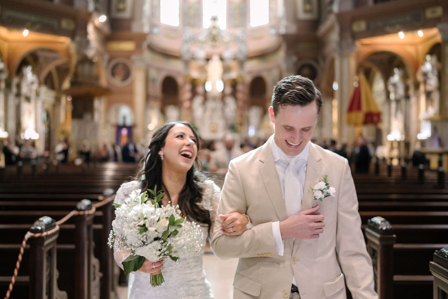 A joyful couple walks down the aisle of a grand cathedral after their wedding ceremony holding hands and laughing.