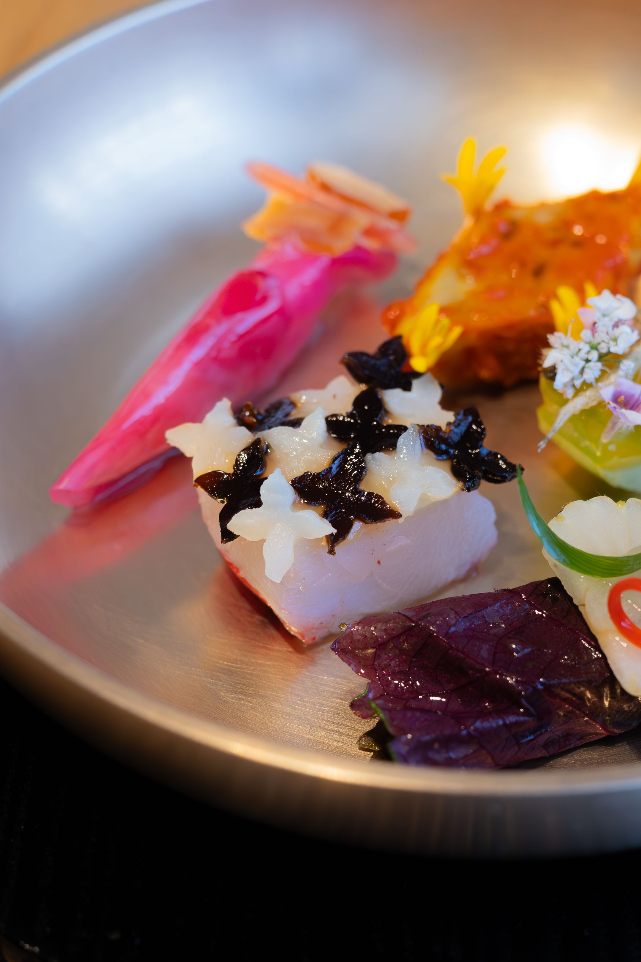 Close-up of artistic sushi pieces garnished with black sauce and edible flowers on a silver plate.