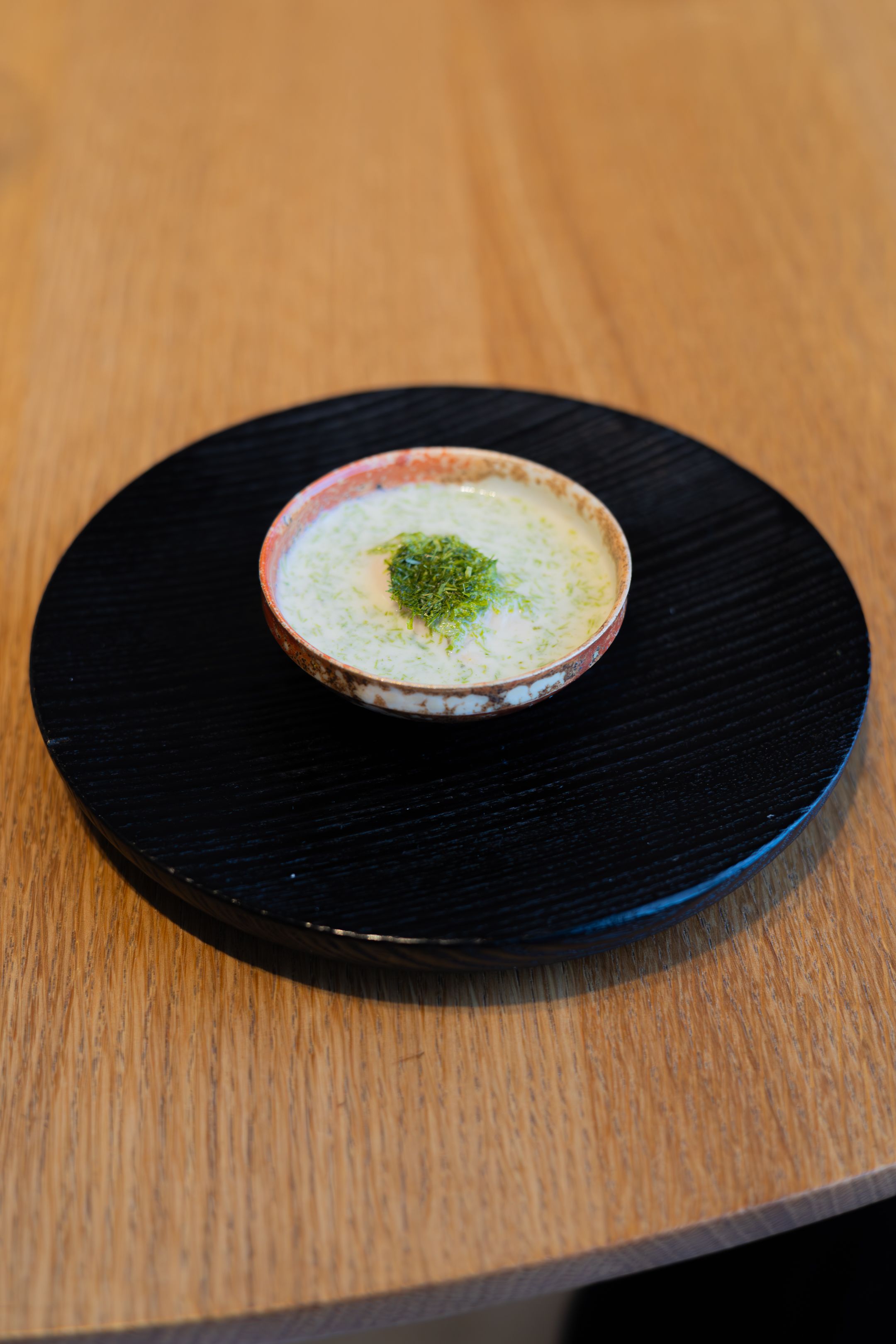 Small ceramic bowl with creamy soup topped with green garnish on a black wooden plate.