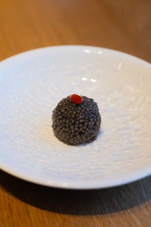 Black spherical truffle with red garnish on white textured plate.
