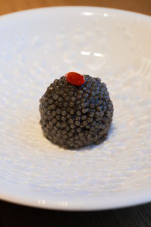 Close-up of a black caviar ball topped with red garnish on white plate.