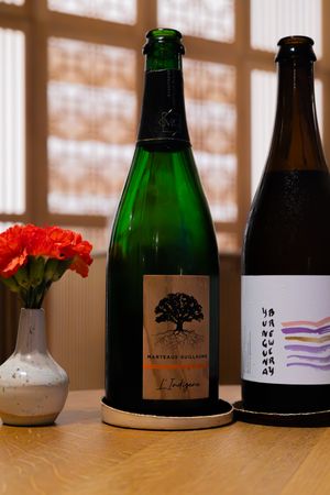 Two wine bottles and red carnations on wooden table with sunlit window reflection.