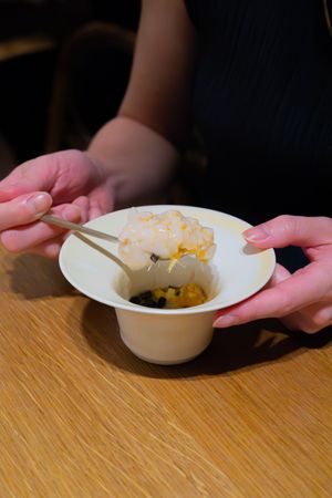 Hands holding a white ceramic bowl containing food with golden broth on a wooden table.