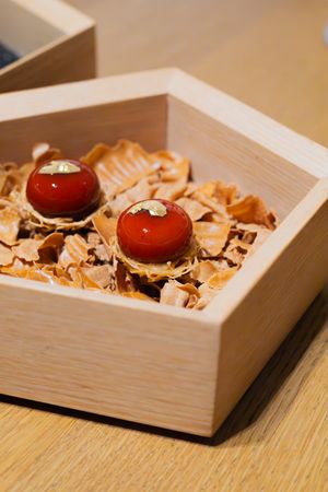 Two red spherical desserts nestled in wood shavings inside a light wooden box.