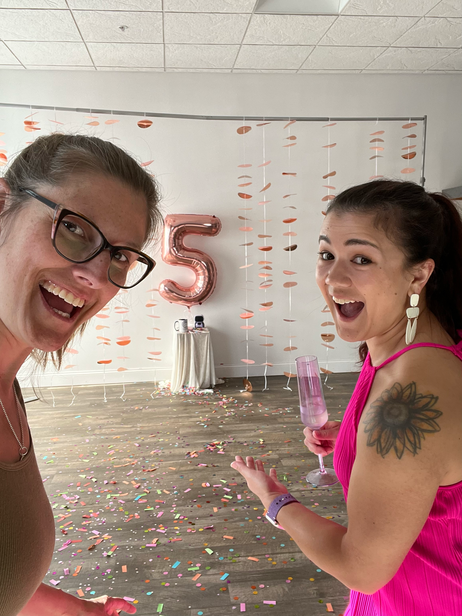 Two people celebrating with champagne and rose gold number 5 balloon amid confetti.