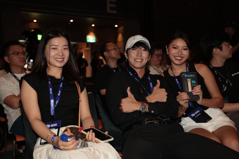 Three conference attendees smile and give thumbs up at a Web3 blockchain event venue.