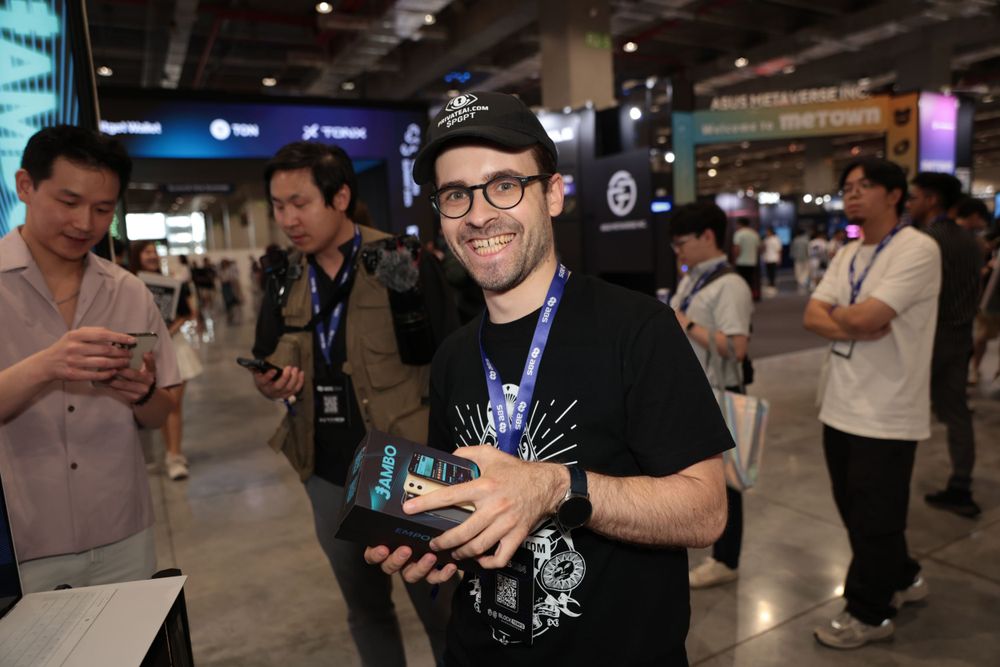 Smiling bearded man holds Jambo Phone product at a busy blockchain conference expo hall.