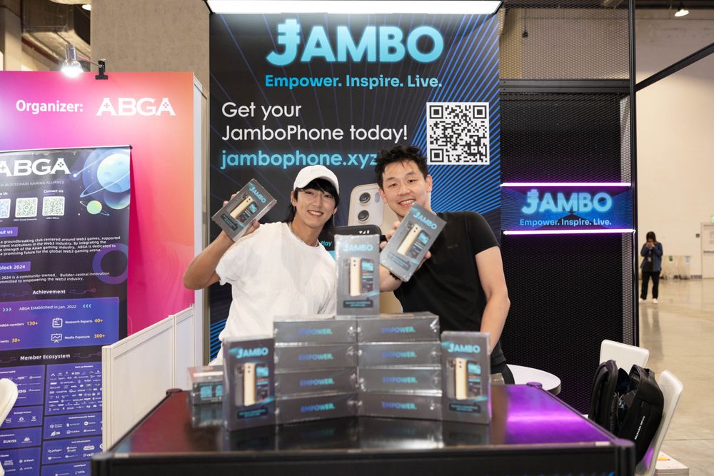 Two people smile at Jambo phone booth displaying products at tech expo.
