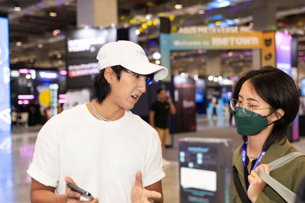 Young man in white cap speaks with masked woman at tech convention.