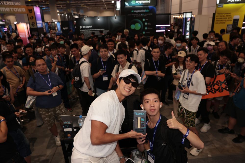 Man poses with fan holding a Jambo phone at a busy tech convention.